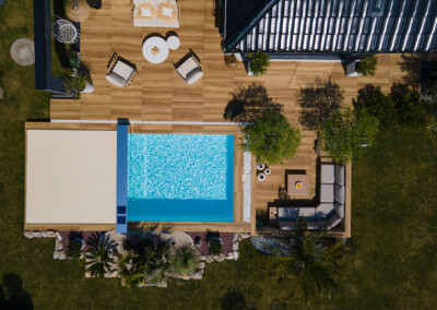 Una piscina azul rodeada de terraza de madera con muebles de exterior y plantas, vista desde arriba.