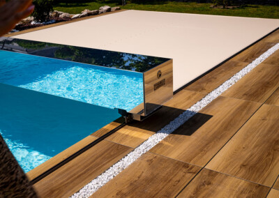 Cubierta automática beige sobre piscina, rodeada de tarimas de madera y piedras blancas decorativas en un jardín.