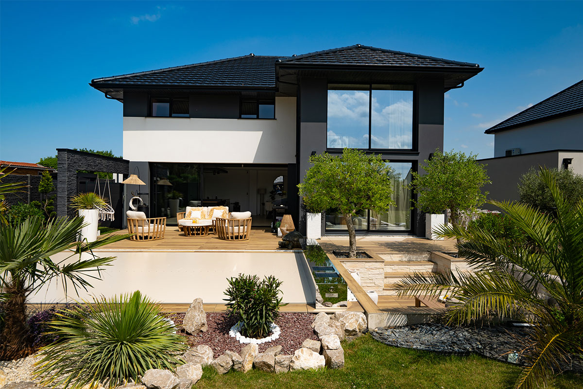 Casa moderna de dos pisos con terraza amueblada y jardín con palmeras y arbustos. Fachada blanca y gris con grandes ventanales.