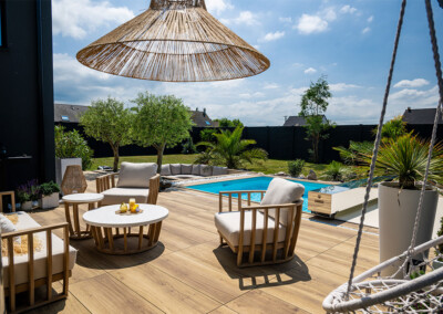 Muebles de patio de madera junto a una piscina, rodeados de plantas verdes y un cielo azul despejado.