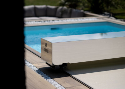 Cubierta automática de piscina de acero inoxidable junto a una piscina azul rodeada de piedras blancas y suelos de madera clara.