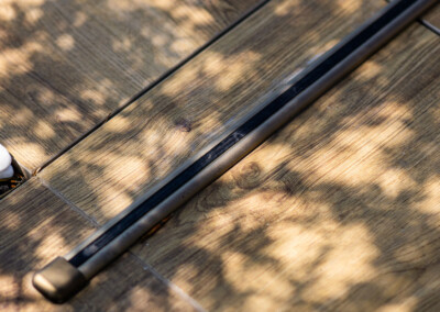 Una cubierta de madera con sombras de hojas y un riel metálico oscuro en primer plano.